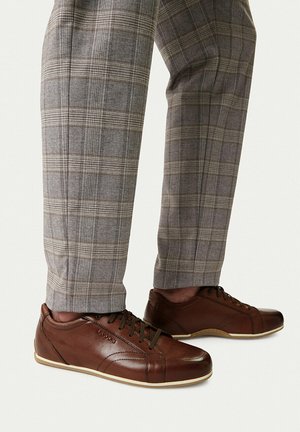 Scarpe stringate in pelle marrone indossate con pantaloni grigi a quadri su sfondo bianco.