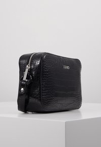 Černá kabelka z umělé krokodýlí kůže přes rameno s trhacími zipy a logem značky na přední straně, umístěná na bílém podstavci.