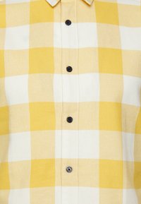 Camisa de cuadros de manga corta con un patrón a cuadros amarillo y blanco, con cierre de botones en el frente y botones negros. Tela de algodón texturizada.