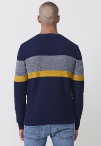 Marineblaues Sweater mit gerippter Textur, der horizontale Streifen in Grau und Senfgelb über die Brust und die Ärmel zeigt. Rundhals-Design.