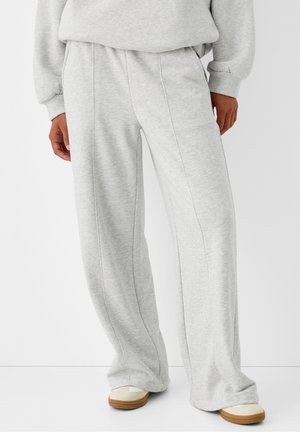Person iført lysegrå vide joggingbukser og matchende sweatshirt, stående med beige sneakers på hvid baggrund.