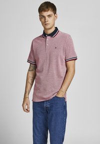 Rød polo-shirt med korte ærmer og knaplukning med tre knapper, marineblå krave og kontrasterende stribede detaljer. Denimjeans fuldender outfittet.