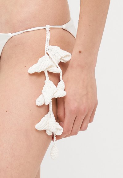 Hvid hæklet blomsterapplikation detaljeret fastgjort til en talje, med et struktureret design og en hængende frynseaccent.