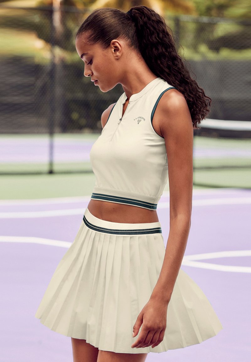 Weißes sportliches Crop-Top mit Reißverschluss und grünen Akzenten, kombiniert mit einem plissierten weißen Rock. Geschmeidige Textur, entworfen für Sportbekleidung.