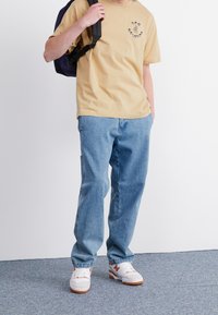 T-shirt en coton beige avec un logo, jean denim bleu et baskets blanches avec des accents orange. Modèle portant un sac à dos noir.