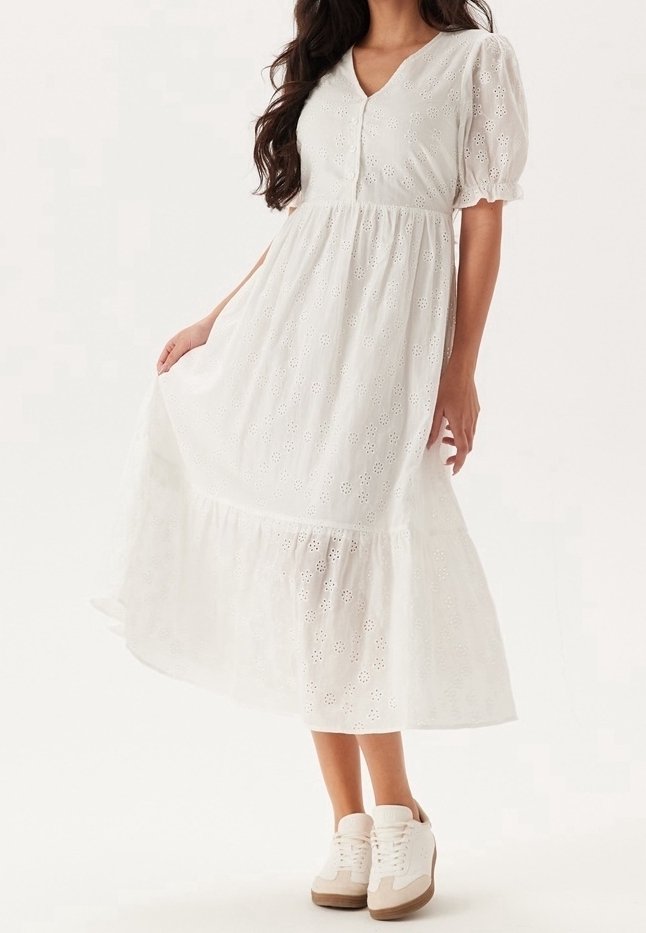 Biała bawełniana sukienka z krótkimi bufiastymi rękawami, dekoltem w kształcie litery V, wzorem ażurowym, warstwowym dołem i koronkowymi detalami, zestawiona z białymi i jasnobrązowymi trampkami.