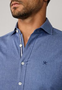 Chemise en chambray bleu avec un col boutonné, ornée de boutons blancs, d'accents rayés navy à l'intérieur du col et d'un logo brodé sur la poitrine.