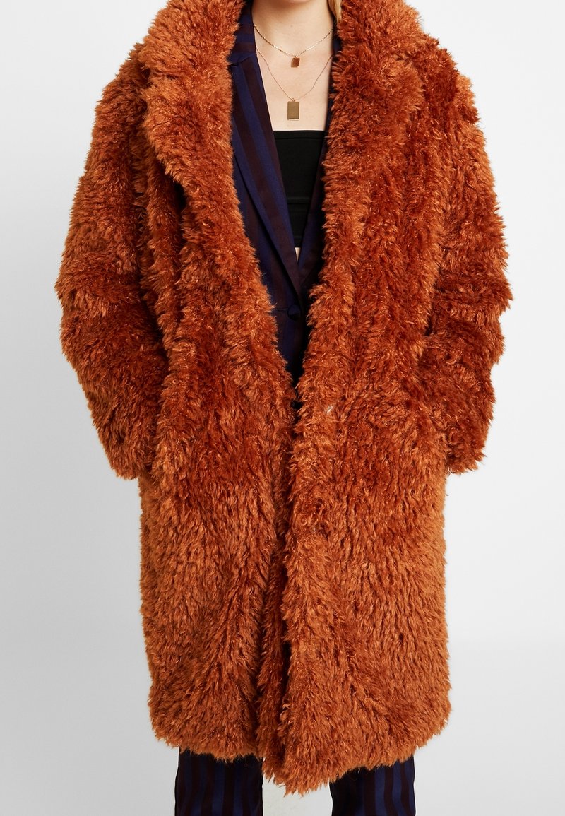 Long manteau en fausse fourrure couleur rouille porté sur un blazer rayé foncé et un haut noir, mains du modèle dans les poches du manteau, vue de face.