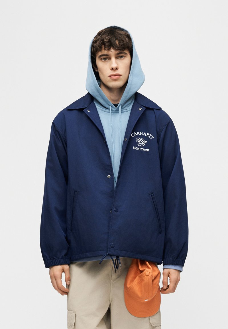 Giovane uomo con indosso una felpa con cappuccio blu e una giacca Carhartt blu navy, che tiene in mano un cappellino arancione, in piedi davanti a uno sfondo chiaro e uniforme.