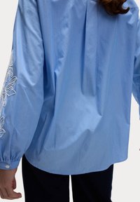 Blusa a righe di color blu chiaro con maniche lunghe, caratterizzata da dettagli floreali ricamati su una manica, e una vestibilità morbida con polsini abbottonati.