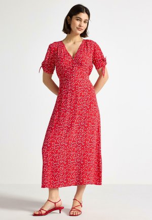 Donna che indossa un vestito midi rosso con pois bianchi e maniche a sbuffo, abbinato a sandali rossi con tacco basso e cinturini, in piedi contro uno sfondo bianco.