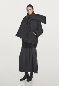 Black quilted jacket with a large scarf; oversized fit, long sleeves, and a structured silhouette. Paired with a long black skirt and boots.