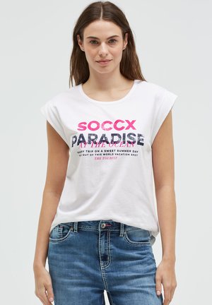 Woman with long brown hair wearing white sleeveless shirt with "SOCCX PARADISE" text and blue jeans standing against a plain light background.