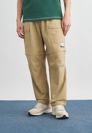 Person wearing beige cargo pants with multiple pockets and beige sneakers, standing on blue carpet.