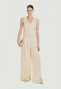 Nainen, joka on pukeutunut beigeen hihattomaan nappivestiin ja leveälahkeisiin housuihin, pitää kädessään pientä teksturoitua clutch-laukkua.