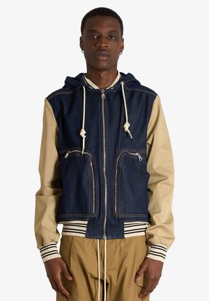 Giovane uomo che indossa un gilet di jeans blu navy con cappuccio sopra maniche beige con polsini a righe e pantaloni beige abbinati, in piedi davanti a uno sfondo semplice.