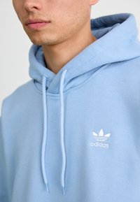 Lyseblå Adidas hettegenser med myk tekstur, med en hvit logo på brystet og hette med snor. Nærbilde av designdetaljene.