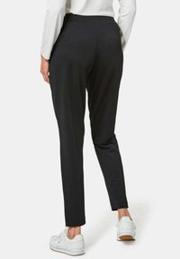 Zwarte tapered broek met een gladde textuur en een elastische tailleband, gecombineerd met witte sneakers. Het uitzicht toont de achterkant en het zijprofiel.
