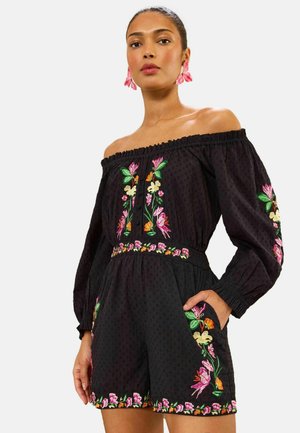 Vrouw draagt zwarte off-shoulder romper met kleurrijke bloemrijke borduursels op mouwen, borst, taille en shorts, handen in zakken, neutrale achtergrond.