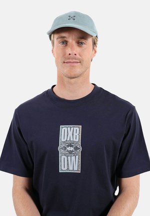 Jeune homme portant une casquette bleu clair et un t-shirt bleu marine avec un graphisme blanc OXB OW centré sur la poitrine, fond neutre.