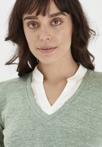 Grøn v-hals sweater med en textured finish, lagd oven på en hvid skjorte med krave, synlige knapper og et glat stof.
