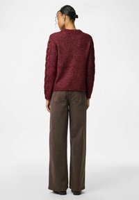 Tumši sarkans adīts džemperis ar teksturētu rakstu un ribu aprocēm, kombinēts ar brīviem brūniem korduroja biksēm, redzams no aizmugures.