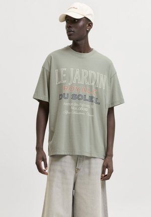 Mężczyzna w oversize zielonym t-shircie z francuskim napisem, szerokich jasnych dżinsach i beżowej czapce z daszkiem, patrzy w bok na białym tle.