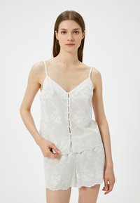 Valkoinen ilman hihoja oleva blouse, jossa on kukkakirjontaa ja nappikiinnitys, yhdistettynä samankaltaisiin shortseihin, joissa on aallokkuureunat. Kevyt kangas.