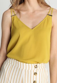 Gul ärmlös topp med V-ringning och metallaccentband, i kombination med vita högmidjade byxor med vertikala ränder som har knappar.