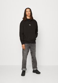Sudadera negra con capucha y bolsillo frontal, presenta un pequeño logo. Combinada con jeans de mezclilla grises y zapatillas negras, de pie sobre un fondo neutro.