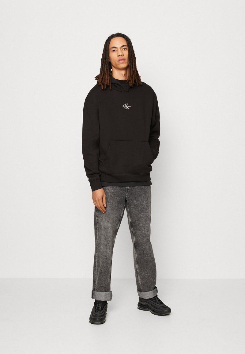Sudadera negra con capucha y bolsillo frontal, presenta un pequeño logo. Combinada con jeans de mezclilla grises y zapatillas negras, de pie sobre un fondo neutro.