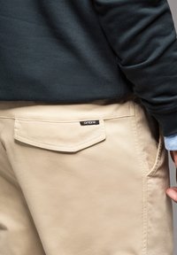 Beige Hose mit einer Gesäßtasche, die einen geschwungenen Klappenverschluss und ein schwarzes Etikett mit weißem Text hat. Begleitet von einem dunklen grünen Sweatshirtärmel.