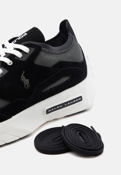 Zapatilla negra y gris con suela y cordones blancos, logo de Polo en el lateral, etiqueta "Ralph Lauren" en la suela, y cordones negros adicionales al lado.