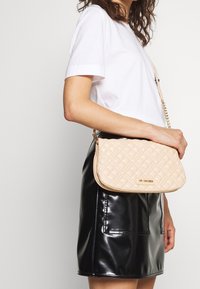 Krémová prošívaná kabelka se zlatými kovovými prvky a řetízkovým popruhem. Hladká koženková textura s pravoúhlým tvarem a jemnými vzory.