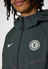 Dark green jacket with a zip closure and hood. Features a white Nike logo and a round Chelsea FC emblem with red accents. Soft texture.