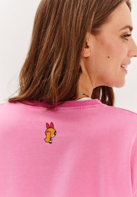 Różowy sweatshirt z małą postacią z kreskówki z blond włosami i czerwonym opaską na plecach. Miękki materiał z okrągłym dekoltem.