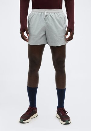 Athlète portant un short de course gris clair, un haut à manches longues bordeaux, des chaussettes bleu marine et des chaussures de course bordeaux, debout devant un fond uni.