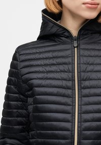 Γυναίκα που φοράει μαύρο μπουφάν με επένδυση, κλεισμένο με φερμουάρ μέχρι το λαιμό, με κουκούλα και οριζόντια ραφές.