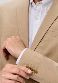 Beige blazermed et struktureret stof, med en enkelt brystlomme og fire brune knapper på manchetterne, båret over en hvid skjorte.
