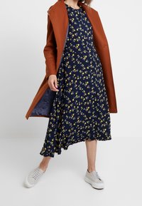 Vestido midi floral en navy con acentos amarillos, combinado con un abrigo largo color óxido. Presenta una tela suave y dobladillo con volantes. Terminado con zapatillas blancas.
