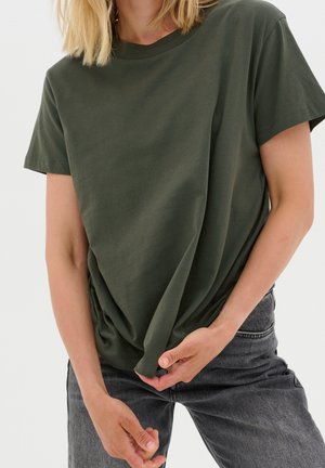 Persoon draagt een donkergroen kortärmelig shirt en grijze jeans, houdt de onderzoom van het shirt met beide handen vast.