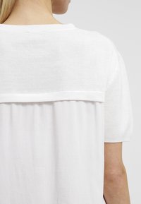 Top bianco lavorato a maglia con inserti testurizzati, tessuto morbido e schiena fluida. Le maniche sono corte e la vestibilità è rilassata.