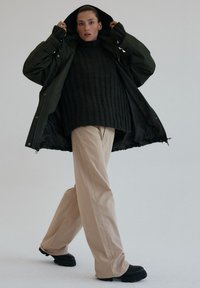 Zielona parka z zamkiem z przodu, nałożona na ciemny, prążkowany sweter, beżowe szerokie spodnie i czarne masywne buty.