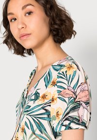 Blusa floral com uma mistura de flores cor-de-rosa e amarelas sobre um fundo creme, com folhas verdes e mangas curtas em balão.