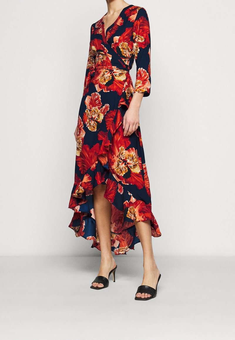 Robe portefeuille fleurie à manches longues, fond bleu marine, fleurs rouges et orange, ourlet irrégulier avec bordure à volants ; portée avec des talons noirs.