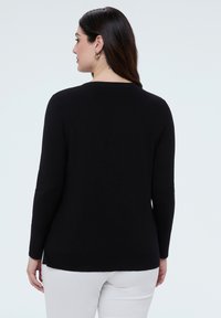 Frau mit langen dunklen Haaren, die ein schwarzes Langarmoberteil und weiße Hosen trägt, von hinten vor einem schlichten hellen Hintergrund.