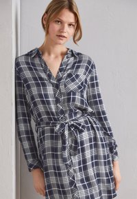 Abito camicia a scacchi navy e bianchi con maniche lunghe, abbottonatura frontale, colletto, tasca sul petto e cintura legata in vita. Tessuto dalla texture liscia.