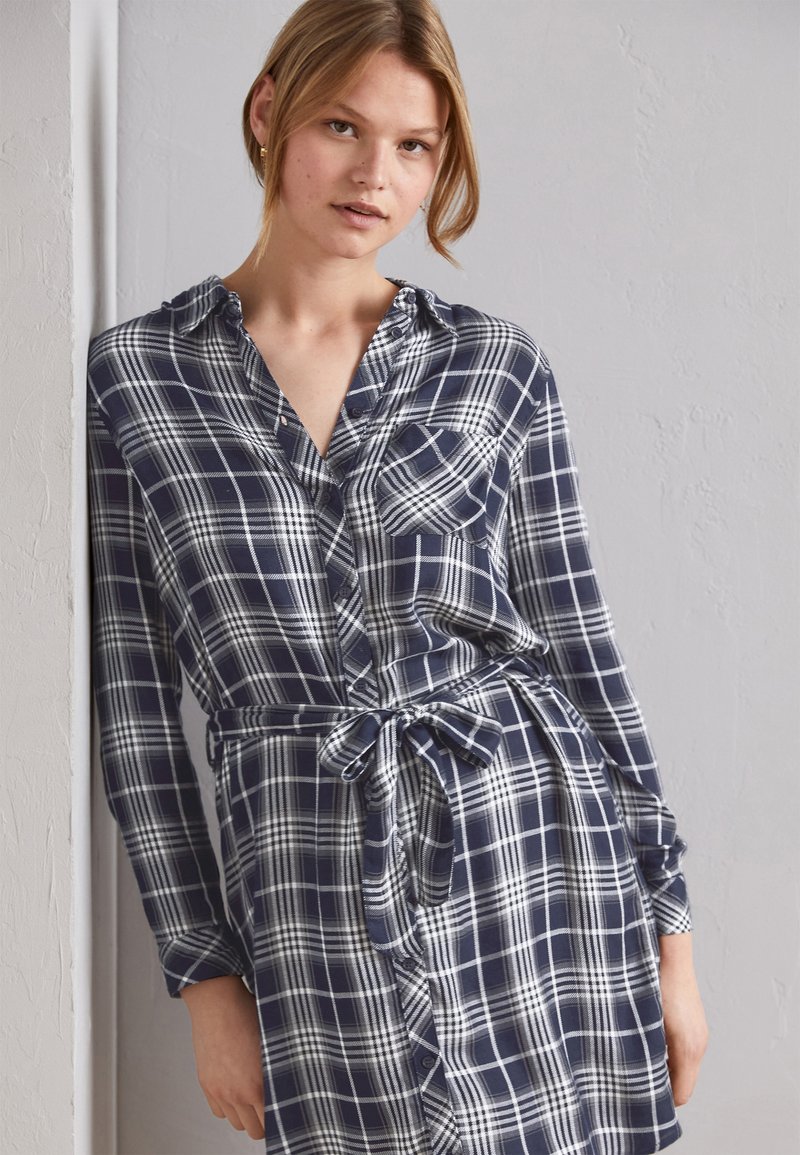 Abito camicia a scacchi navy e bianchi con maniche lunghe, abbottonatura frontale, colletto, tasca sul petto e cintura legata in vita. Tessuto dalla texture liscia.