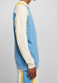 Vista laterale di una persona che indossa una camicia a maniche lunghe blu e crema con accenti gialli e pantaloni abbinati blu, crema e giallo.