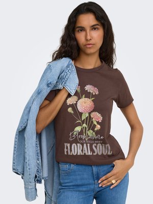 Mujer con cabello largo y oscuro que lleva una camiseta marrón con flores rosas y texto, sujetando una chaqueta vaquera azul sobre su hombro.